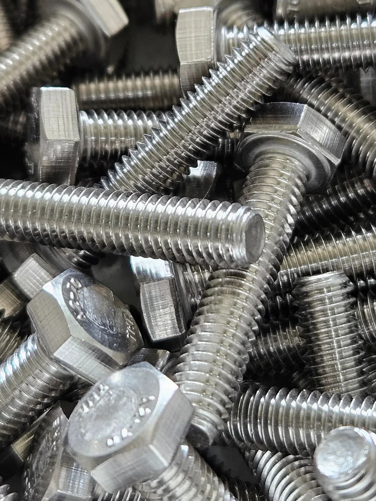 A pile of hex head bolts and threaded studs in stainless steel, plain finish, with grade markings stamped on the heads.