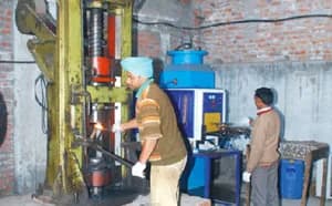 Hot forging: an operator holds a heated part at a forging press while a second worker stands at the machine beside it.